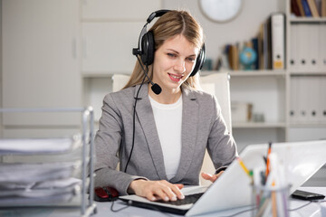 Young positive woman customer support phone operator in microphone headset working on laptop in the...