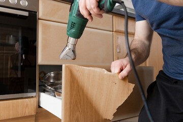 A man repairs a kitchen cabinet using a heat gun to remove old veneer or glue. DIY home improvement and renovation concept, showing how people can improve their living space with their own hands.