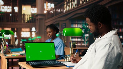 Med school student works on his thesis next to isolated mockup display in the library, doing medical research and reviewing specialty books. Future doctor studies with dedication. Camera A.