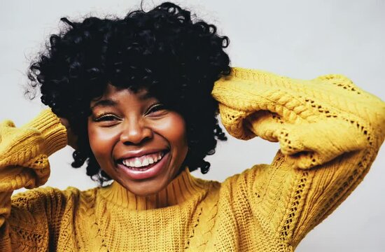 A cheerful moment. Closeup portrait of smiling black woman with hands on curly hair looking up wearing a yellow sweater isolated against a gray background