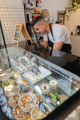Smiling bakery owner in a black apron leans over the glass counter, adjusting packaged food. Fresh meals and pastries, menu labels, modern shop interior, warm ambiance, customer service