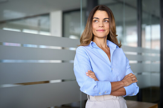 Smiling successful mature businesswoman of middle age standing in office arms crossed. Happy confident older professional business woman leader executive looking away thinking of future success.
