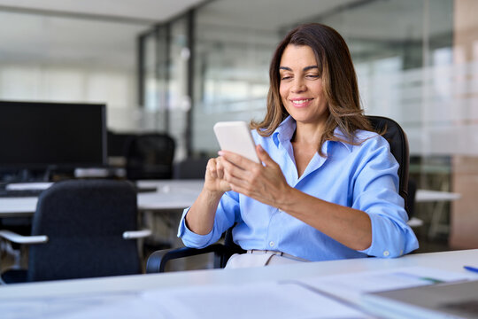 Smiling mature business woman executive using cell phone working in office. Happy middle aged businesswoman holding smartphone, professional female executive looking at cellphone at workplace.