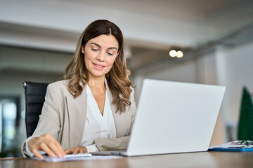 Happy middle aged 45 years old business woman professional hr manager reading cv or finance report overview, lawyer attorney checking document, working on laptop computer sitting at desk in office.