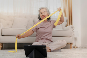 An elderly woman exercises with a resistance band at home, focusing on health and fitness. She...