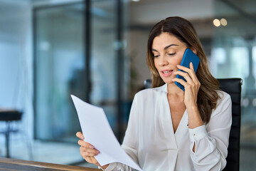 Busy middle aged professional business woman executive making call having conversation at work. Mature female manager or entrepreneur talking on the phone checking document sitting at office desk.
