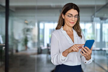 Smiling busy middle aged business woman executive wearing eyeglasses using smartphone in office. Happy mature female employee or professional bank manager looking at cell mobile phone tech at work.