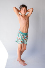 Young preteen beach boy wearing swim trunks standing with his hands behind his head with serious expression © 3Days2Go Media