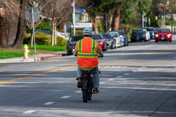 One cyclist wearing a bright orange safety vest and helmet rides an electric bike along a marked urban street, surrounded by parked cars and lush greenery. Rear view.