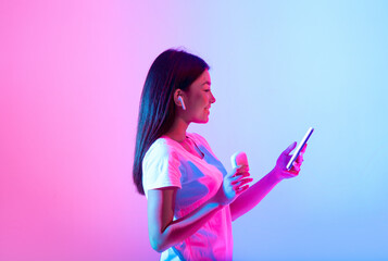 A young woman with long hair holds a smartphone and a wireless earbud, smiling as she listens to...