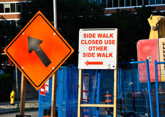 Obraz premium Bright orange and white detour signs at a construction site direct pedestrians to use an alternate sidewalk. Blue safety fencing and heavy machinery are visible in the background.