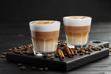 Tasty latte macchiato in glasses, coffee beans and cinnamon on black wooden table, closeup