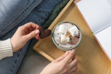 Woman having tasty hot chocolate with cookie while reading book, top view