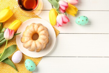 Delicious bundt cake, Easter eggs, tea and tulips on white wooden table, flat lay. Space for text