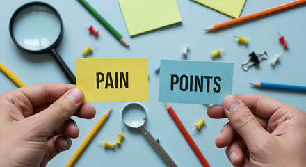 Hands holding cards labeled 'PAIN' and 'POINTS,' surrounded by office stationery like push pins, pencils, and a magnifying glass, symbolizing the process of identifying challenges