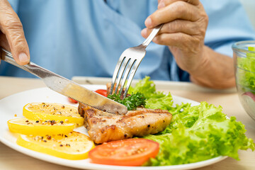 Asian senior woman patient eating pork chop stake and vegetable salad for healthy food in hospital.