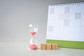 Hourglass sand timer with three blank wooden toy blocks next to a calendar, on a white surface.