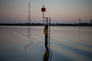 A navigational marker with a caution sign stands in calm water at dusk, its reflection distorted by gentle ripples, with power lines and an industrial backdrop