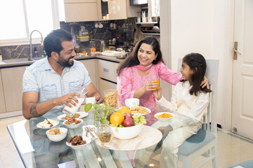 indian mother giving orange juice to her daughter in breakfast. young indian family of three having morning meal at home.