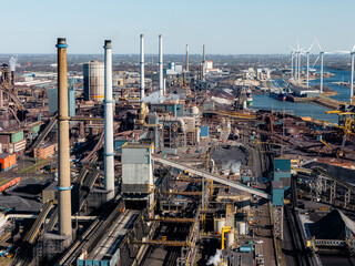Industrial steel plant in IJmuiden, Netherlands, with large chimneys, conveyor belts, and emissions. Aerial view of a major production hub, showing both traditional and renewable energy elements.