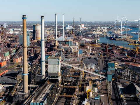 Industrial steel plant in IJmuiden, Netherlands, with large chimneys, conveyor belts, and emissions. Aerial view of a major production hub, showing both traditional and renewable energy elements.