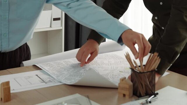 Smart architect and safety engineer discussion about building blueprint on table with architectural document, measurement tools and wooden block scatter around at modern office. Closeup. Delineation.
