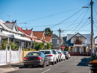 A residential street in Australia, lined with old suburban houses in an inner suburb, with overhead power lines connecting historic homes. Cars are parked along the street.