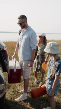 Vertical shot of family walking to car, loading belongings into trunk while leaving sunny lakeshore after camping trip