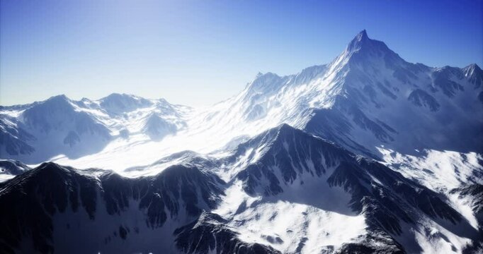Snow blankets the rugged peaks and valleys of a mountain range, illuminated by bright sunlight. The clear blue sky enhances the beauty of the serene, chilly landscape.