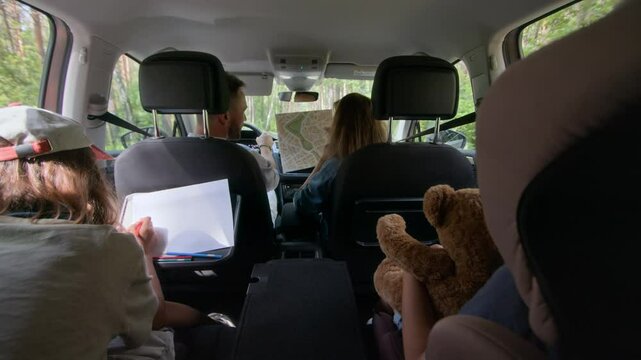 View from backseat of children drawing on paper and holding stuffed toy while parents discussing route on paper map and driving during family road trip in woods