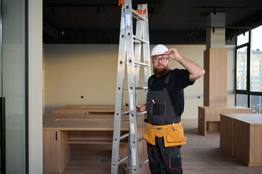 Construction worker holding ladder. Man carrying steel ladder. Safe at work. Bearded builder standing in warehouse wearing hardhat. Repairs concept. Construction builder worker carrying ladder