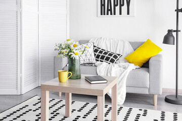 Interior of stylish living room and vase with blooming chamomile flowers on wooden table