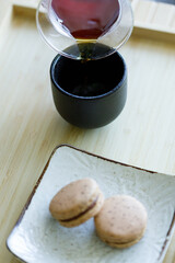 Black coffee pouring into cup and black tea flavor macaron on wooden background.