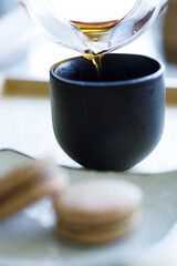 Black coffee pouring into cup and black tea flavor macaron on wooden background.