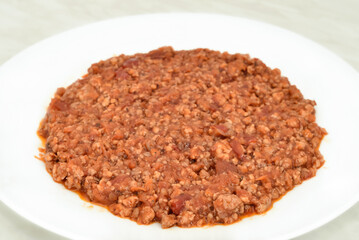 Stewed mixed pork and beef minced meat with peeled and chopped tomatoes in tomato juice on the white plate on the light gray marble table texture. Close-up. Selective focus.