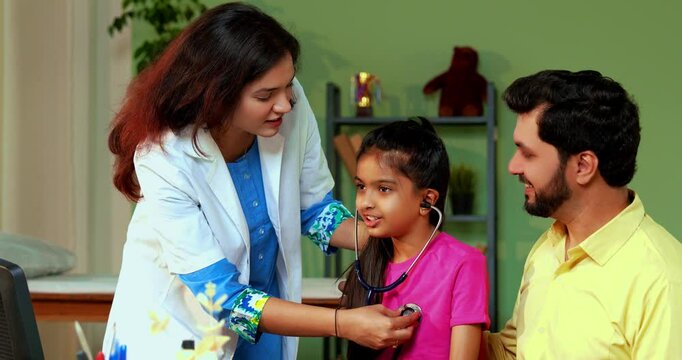 Indian Asian playful female doctor or pediatrician engaging young girl patient by letting her hear own heartbeat using stethoscope in a warm and friendly clinic atmosphere