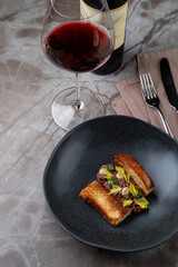 An exquisite dish is presented on a sleek black plate, garnished creatively. A glass of rich red wine stands beside it, enhancing the sophisticated dining experience in a stylish restaurant
