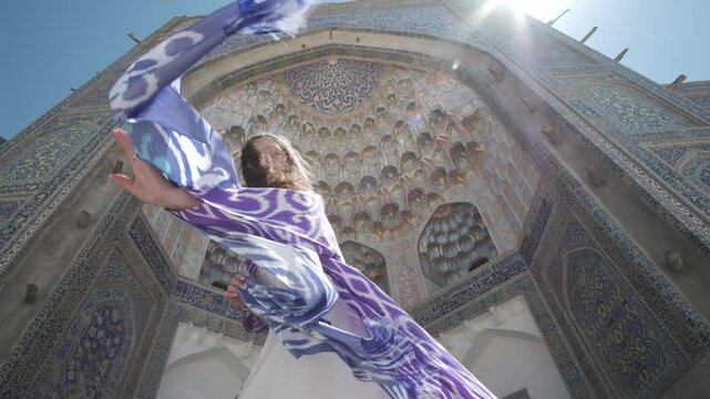 A young woman with long hair in an oriental robe and skullcap stands in the wind near an ancient mosque
