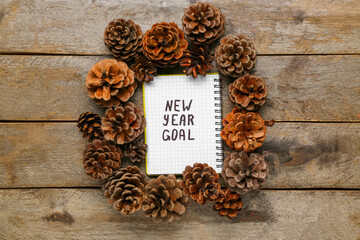 Notebook with text NEW YEAR GOAL and pine cones on wooden background