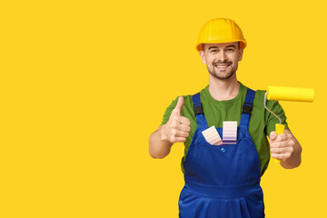 Portrait of male painter with color palettes and paint roller showing thumb-up on yellow background