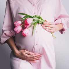 Pregnancy, Motherhood, Mother's Day Holiday concept. Young woman in maternity shirt dress with tulips flowers holds hands on belly. Beautiful pregnant woman waiting for baby birth.