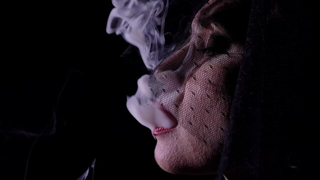 Retro woman actress with pin-up hairstyle and makeup isolated on black background, wearing veil and smoking mouthpiece cigarette, blows out smoke. Slow motion.