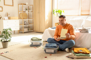 Young man with notebook folding charity clothes in bedroom