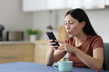 Fototapeta premium Surprised asian woman using smart phone in a kitchen