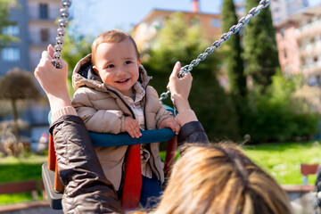 Happy baby boy swinging at the playground with mother pushing