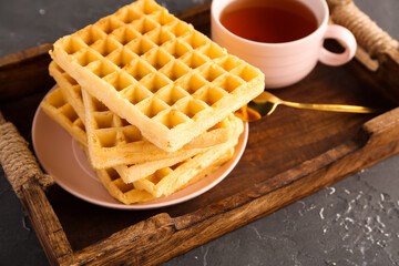 Wooden tray with delicious Belgian waffles and cup of tea on black background
