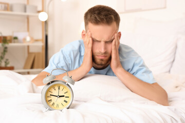 Stressed young man lying on bed with clock at home. Insomnia concept