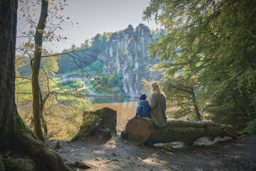 Mom and baby sit on a fallen tree trunk with their backs to the camera and look out over the lake and high cliffs