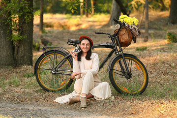 Obraz premium Beautiful young happy woman with bicycle, camera, wicker basket of flowers and plaid sitting in park, outdoors