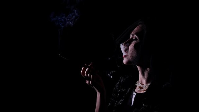 Retro woman actress with pin-up hairstyle and makeup isolated on black background, wearing veil and smoking mouthpiece cigarette, blows out smoke. Slow motion.